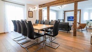A modern dining area with a wooden table and black chairs, a wooden ceiling structure, and a large flat-screen TV in the background.