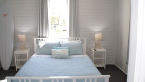 A bedroom with a bed, two bedside tables, a surfboard, and a window with curtains.