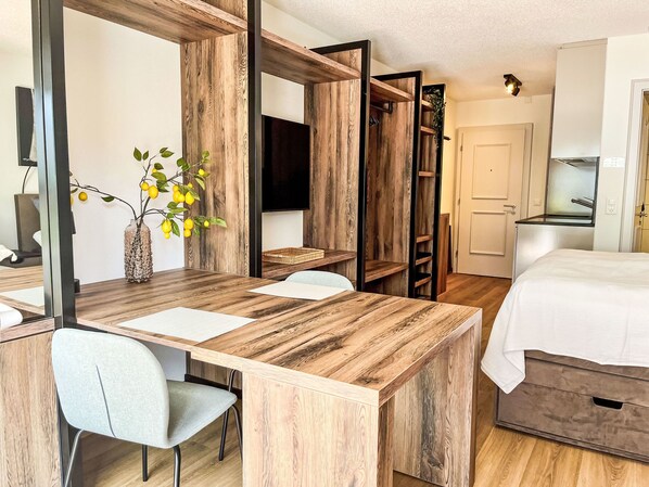 Una habitación moderna con un escritorio de madera, una silla gris, una cama con ropa de cama blanca y un televisor en la pared.