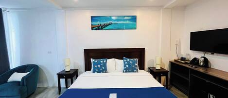 A hotel room with a large bed, a TV, a desk with a phone and coffee maker, and a painting of a beach scene.
