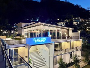 A modern hotel with a prominent sign reading "Porto Novo Anilao." It features a two-story structure with a balcony on the upper level, a staircase leading to the entrance, and a well-lit outdoor area. - Porto Novo Anilao (Mabini)