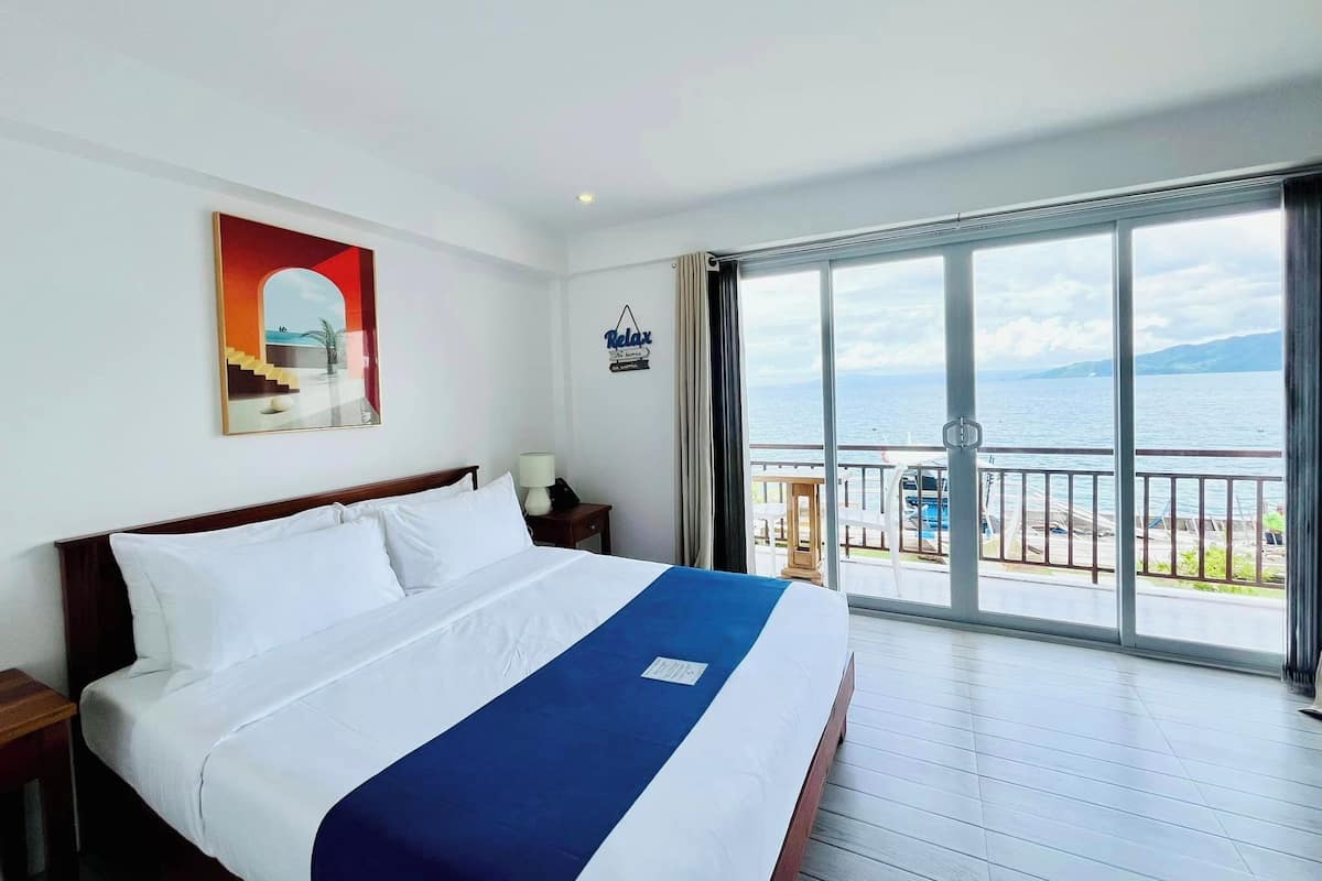 A hotel room with a large bed, a view of the sea, and a balcony with a railing.