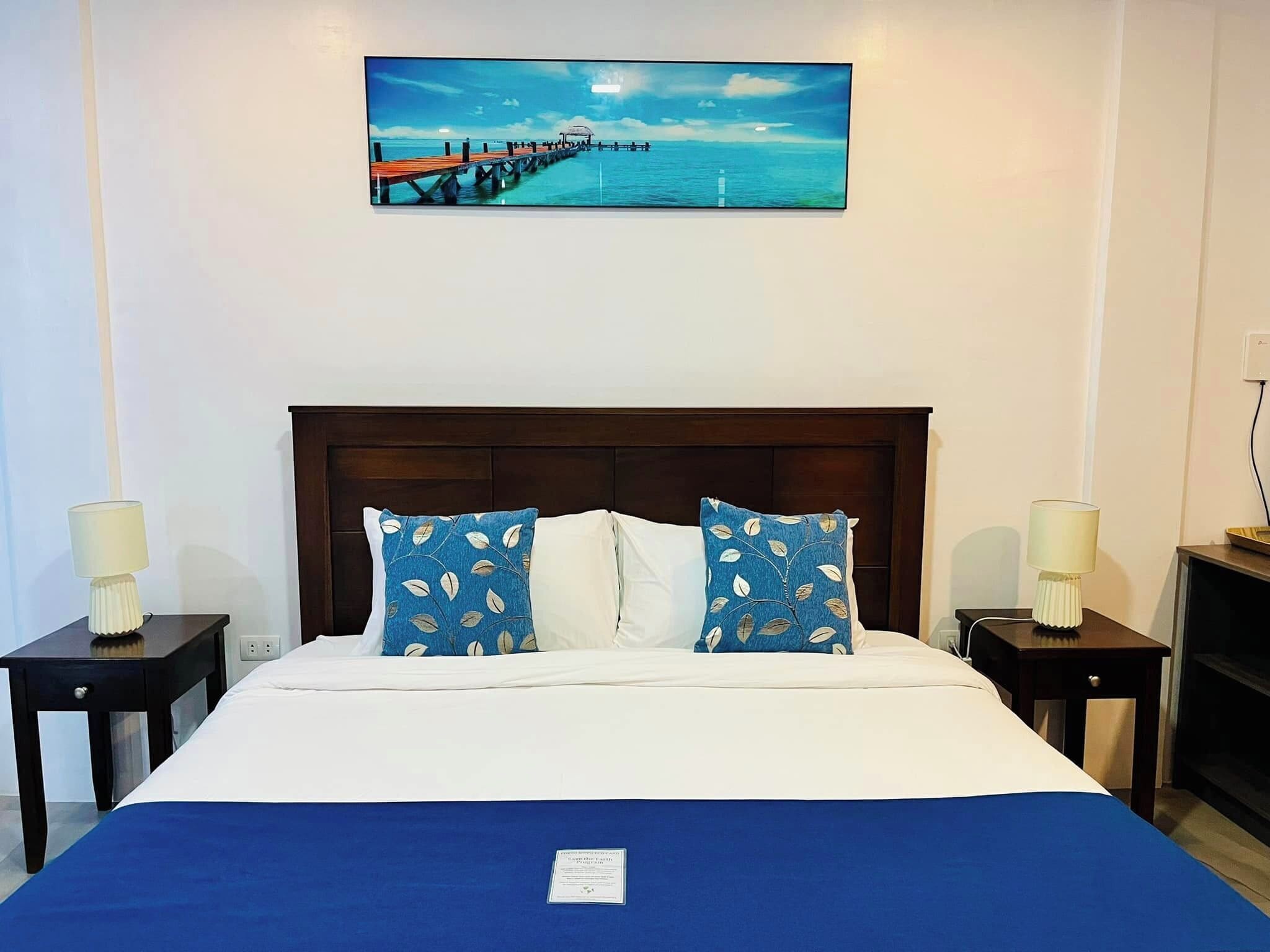 A hotel room with a wooden headboard, a bed with white and blue bedding, two nightstands with lamps, and a painting of a pier over water on the wall.