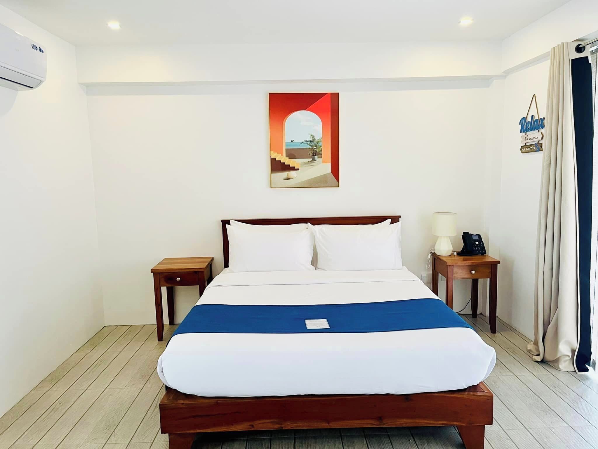 Una cama bien tendida con ropa de cama blanca y azul, un cabecero de madera y una mesita de noche con una lámpara y un teléfono.