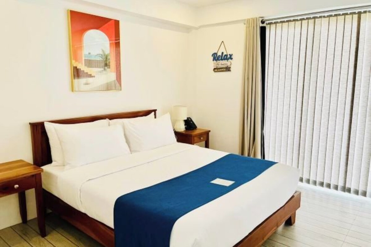 A hotel room with a bed, a nightstand, a lamp, a painting, a sign, and a window with curtains.