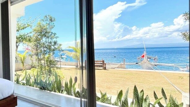 Habitació amb finestra gran que dona a una platja, un llit i vista al mar.