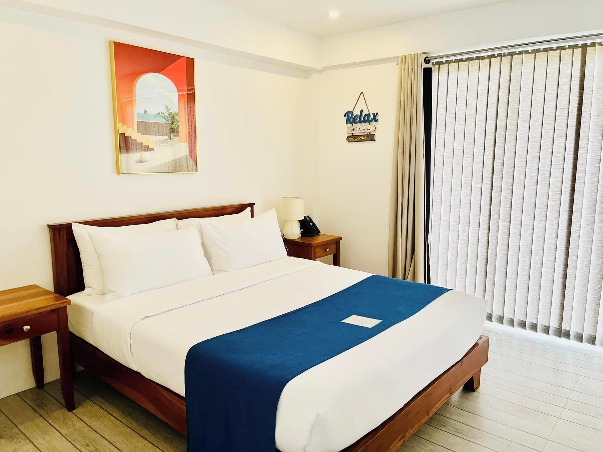 Ein Hotelzimmer mit Bett, Nachttisch, Lampe, Schild und einem Fenster mit Vorhängen.