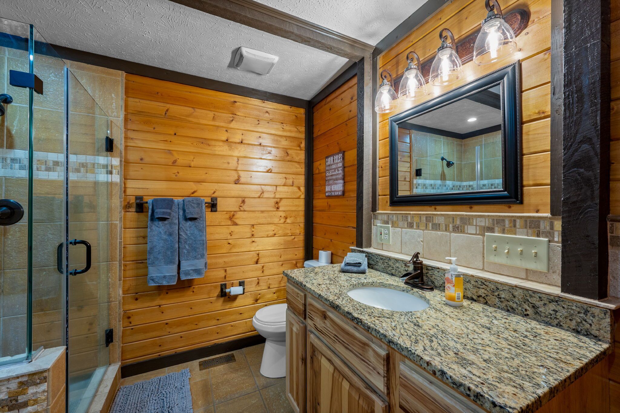 A wooden-paneled bathroom with a glass shower enclosure, a sink, and a mirror with lights.