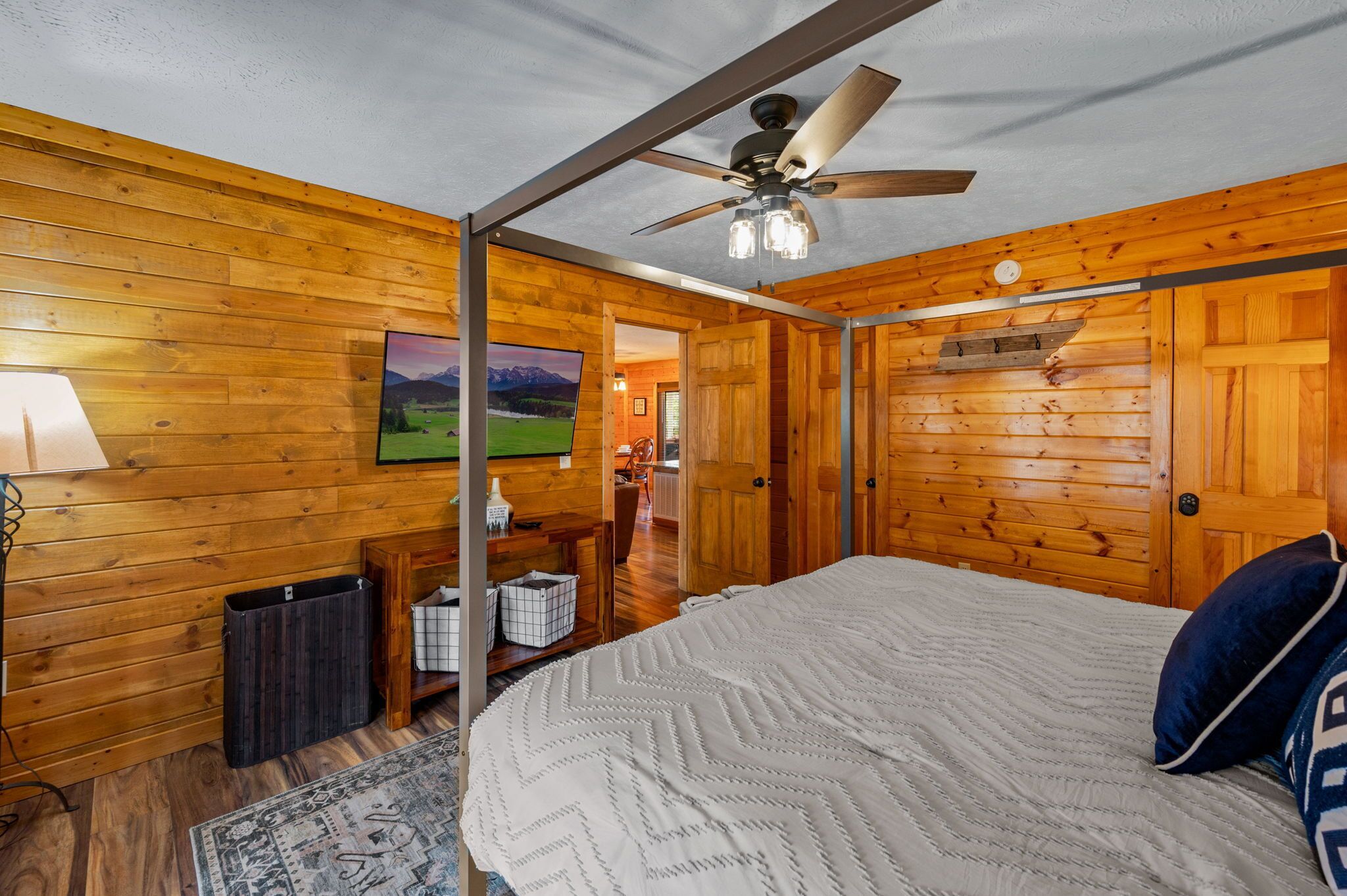 A wooden-paneled room with a bed, a ceiling fan, a framed picture on the wall, and a view into another room.