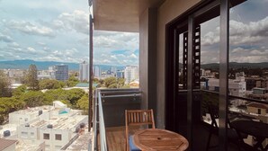 Un balcón con dos sillas de madera y una mesita, con vistas a un paisaje urbano con edificios y árboles.