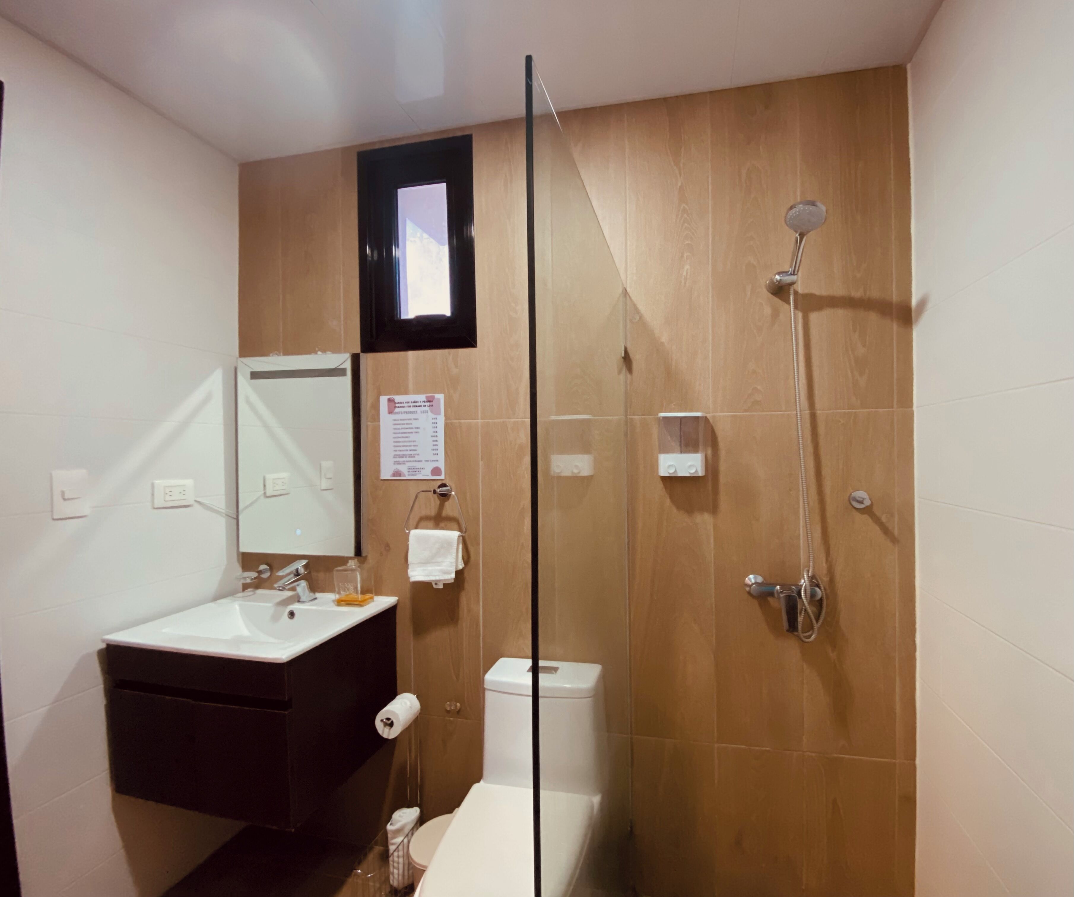 A bathroom with a wooden vanity, a toilet, a glass shower enclosure, and a small window.