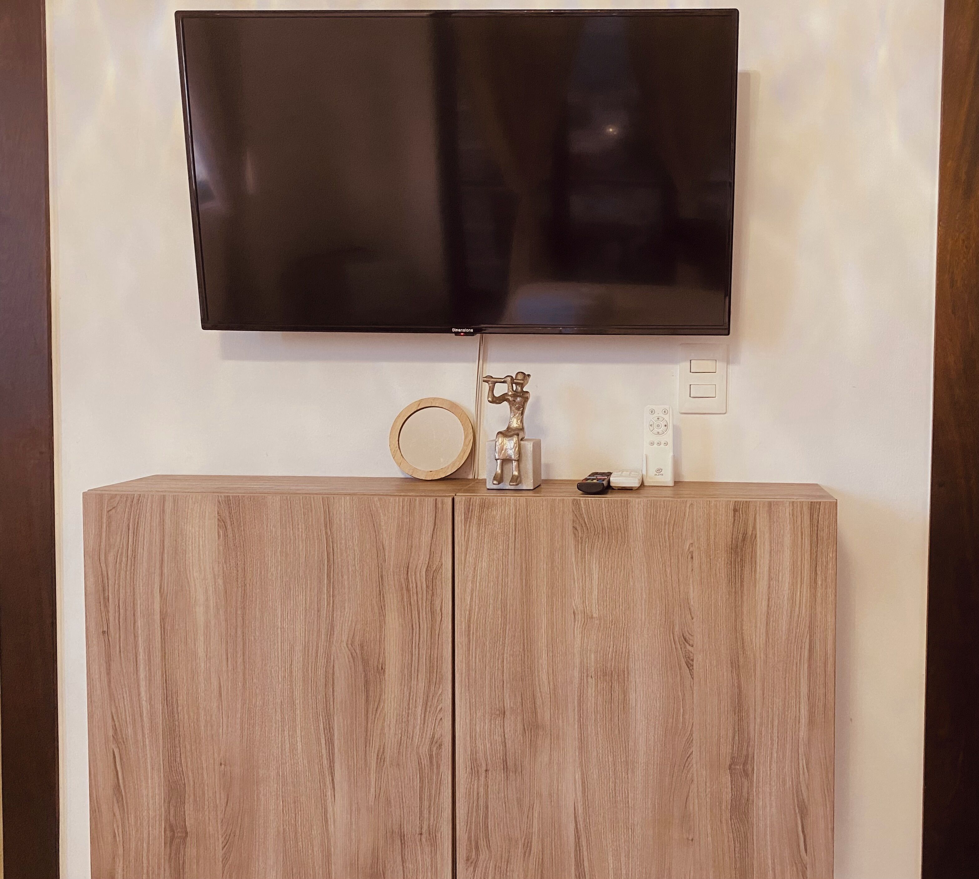 A flat-screen TV mounted on a wall above a wooden cabinet with a round mirror and a small statue.