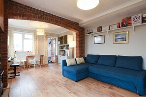 A living room with a blue sectional sofa, a wooden dining table with chairs, a brick fireplace, and a bookshelf with books and decorative items.