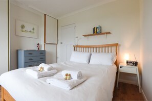 A neatly made bed with white linens and towels, a bedside table with a radio, a blue dresser, and a wall-mounted shelf with books.