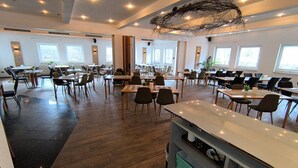 Una àmplia zona de dinar amb diverses taules i cadires, grans finestres i un disseny de sostre modern.