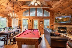 L’intérieur d’un chalet en bois comprend une table de billard, un coin repas, un canapé en cuir, une cheminée et de grandes fenêtres.