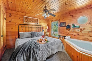Une chambre dans un chalet en bois, avec un lit, un ventilateur de plafond, une fenêtre, une étagère fixée au mur et une baignoire.