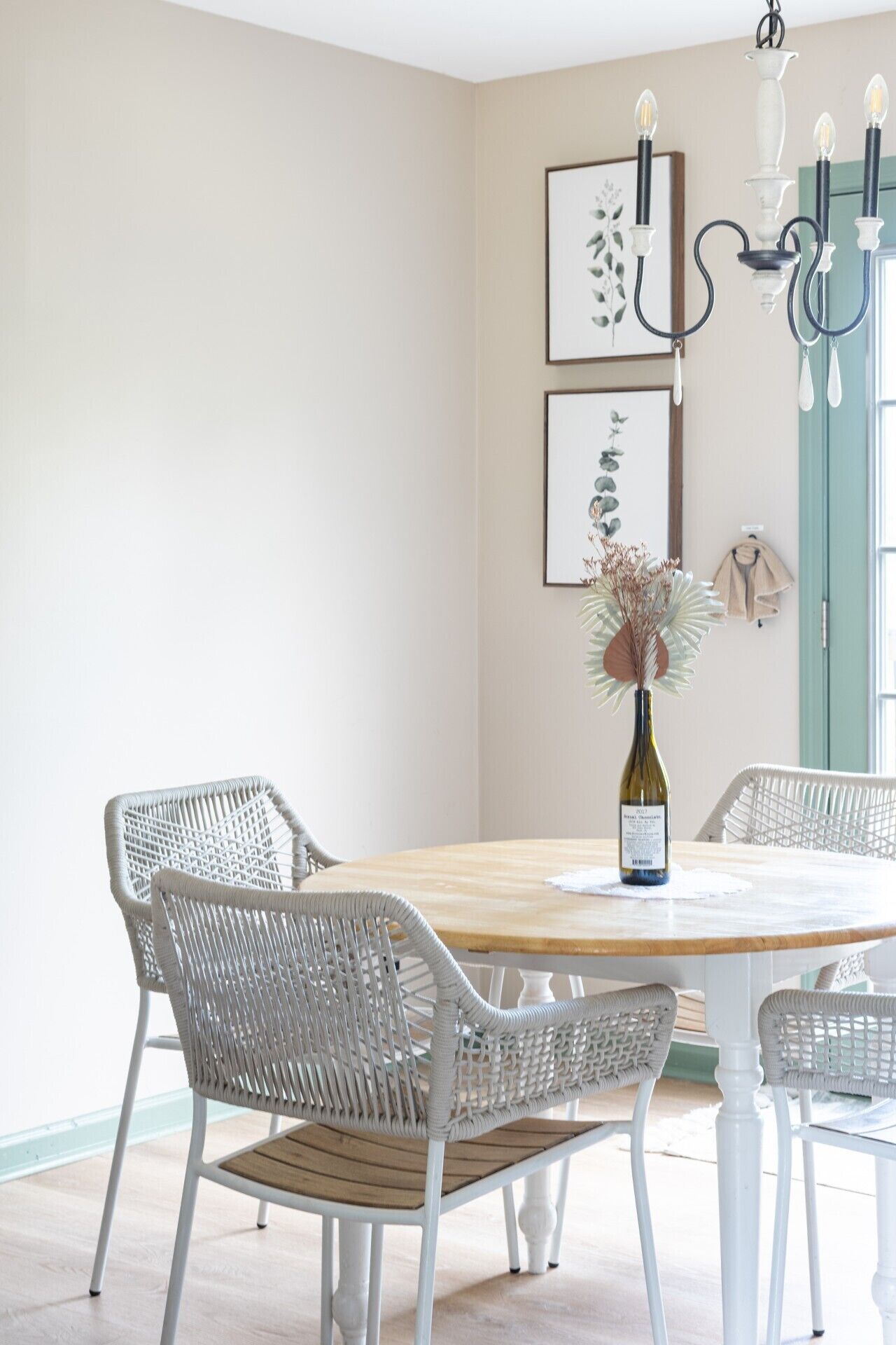 A dining area with a round table, white wicker chairs, a bottle of wine, and framed botanical prints on the wall.