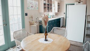 A dining area with a round wooden table, white wicker chairs, a vase with dried flowers, and a kitchen in the background with teal cabinets and a white refrigerator.