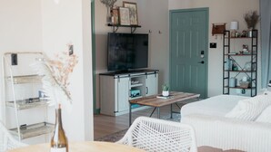 A living room with a white sofa, a coffee table, and a TV. There is a kitchen area with a stove and cabinets.
