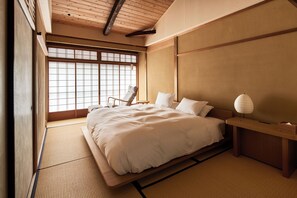 Un dormitorio de estilo japonés tradicional con suelo de tatami, cama baja, mesita de noche de madera y ventana con pantalla shoji.