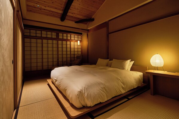 A traditional Japanese-style bedroom with tatami flooring, a low platform bed, a shoji screen, and a bedside table with a paper lantern. - YADORU KYOTO HANARE Kagetsu No Yado (Kyoto)