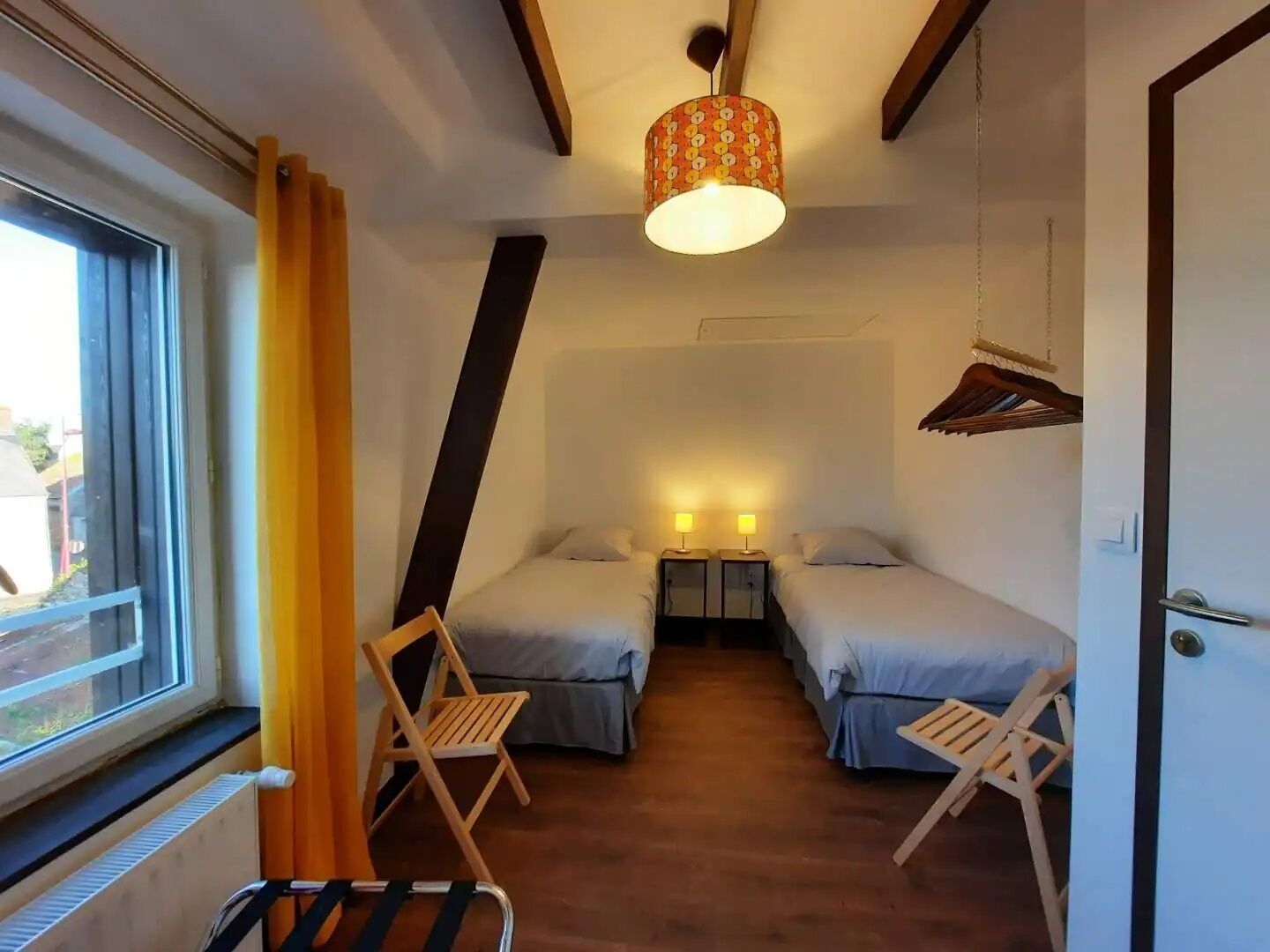 A small, cozy room with two beds, a window with curtains, a hanging light fixture, and a wooden chair.