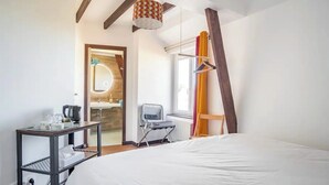 A hotel room with a large bed, a desk with a kettle, a chair, and a window with curtains. - Le petit moulin rouge (Beauvoir)