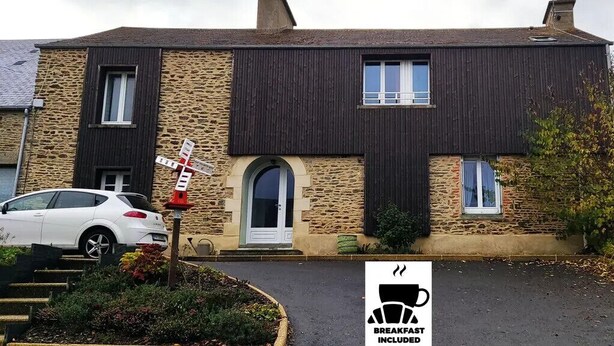 A two-story stone building with a white car parked in front. A sign indicates breakfast is included. - Le petit moulin rouge (Beauvoir)