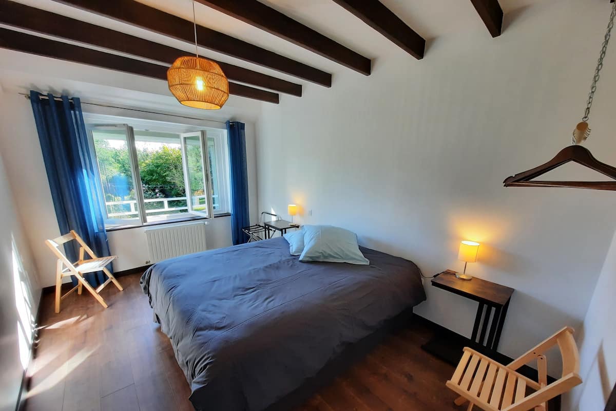 Ein Schlafzimmer mit Bett, Holzboden, einer Hängelampe, einem Fenster mit Vorhängen und einem Nachttisch mit Lampe.
