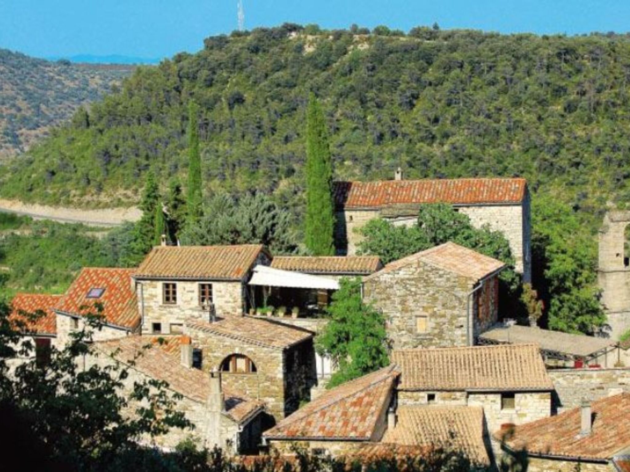 A village with stone buildings and terracotta roofs, surrounded by greenery and hills.