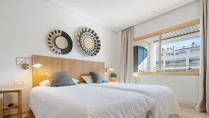 Une chambre d’hôtel moderne avec deux lits, une tête de lit en bois, une grande fenêtre donnant sur des immeubles et deux assiettes décoratives fixées au mur.