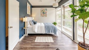 A modern bedroom with a large bed, a nightstand, a floor lamp, and a view of the outdoors through large windows.