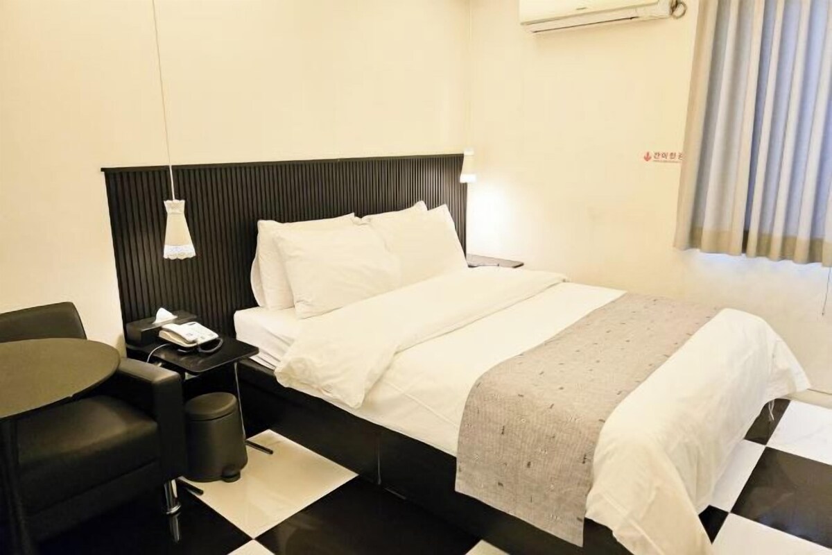A hotel room with a bed, a bedside table, a chair, a small table, and a trash bin.