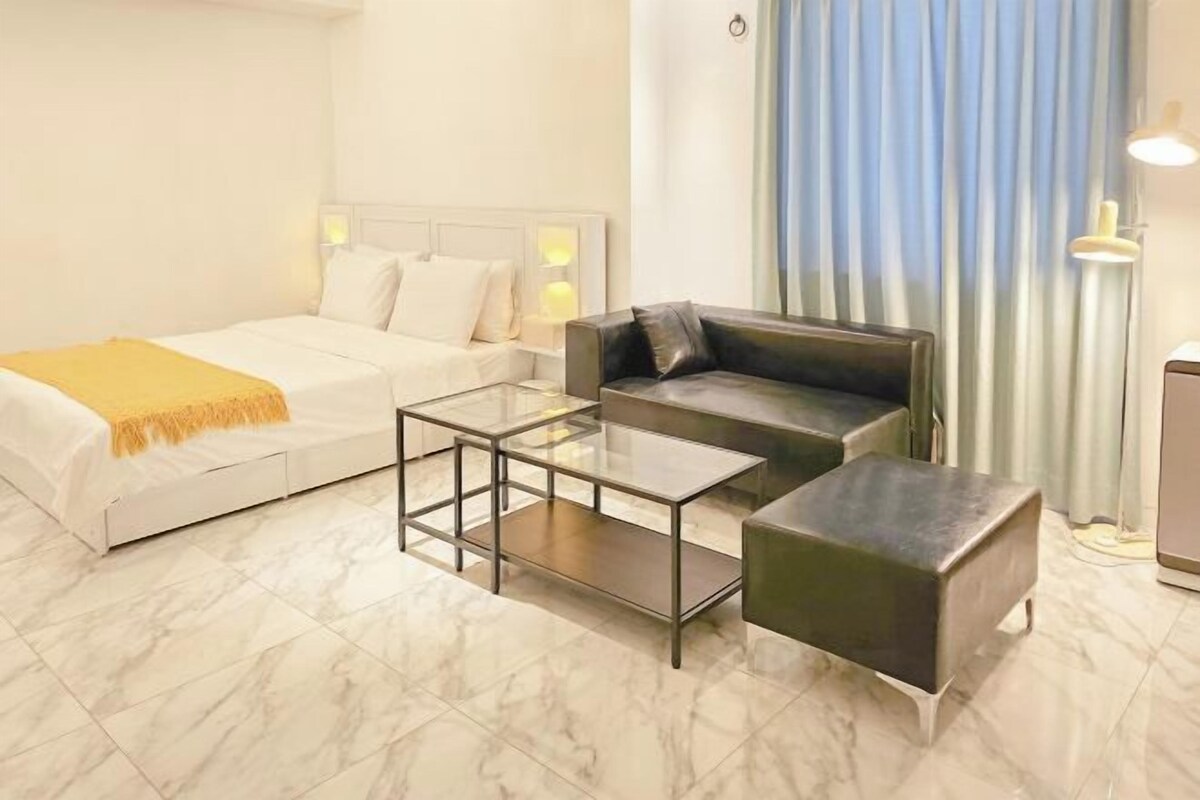 A hotel room with a bed, a sofa, a glass coffee table, and a small ottoman.