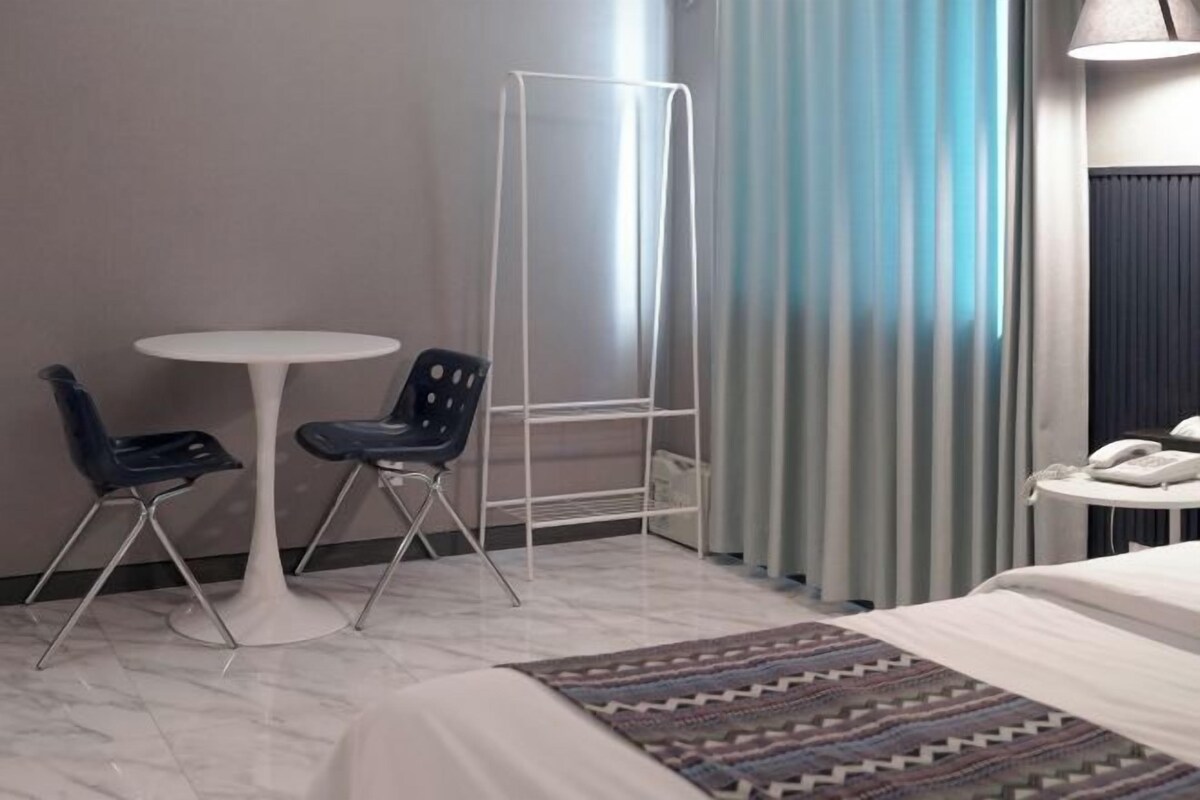 A hotel room with a bed, a small round table, two chairs, a standing shelf, a telephone, and a window with curtains.