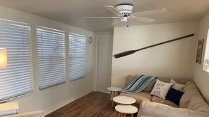 Un salon avec un ventilateur de plafond, un canapé, deux tables d’appoint et une fenêtre avec des stores.