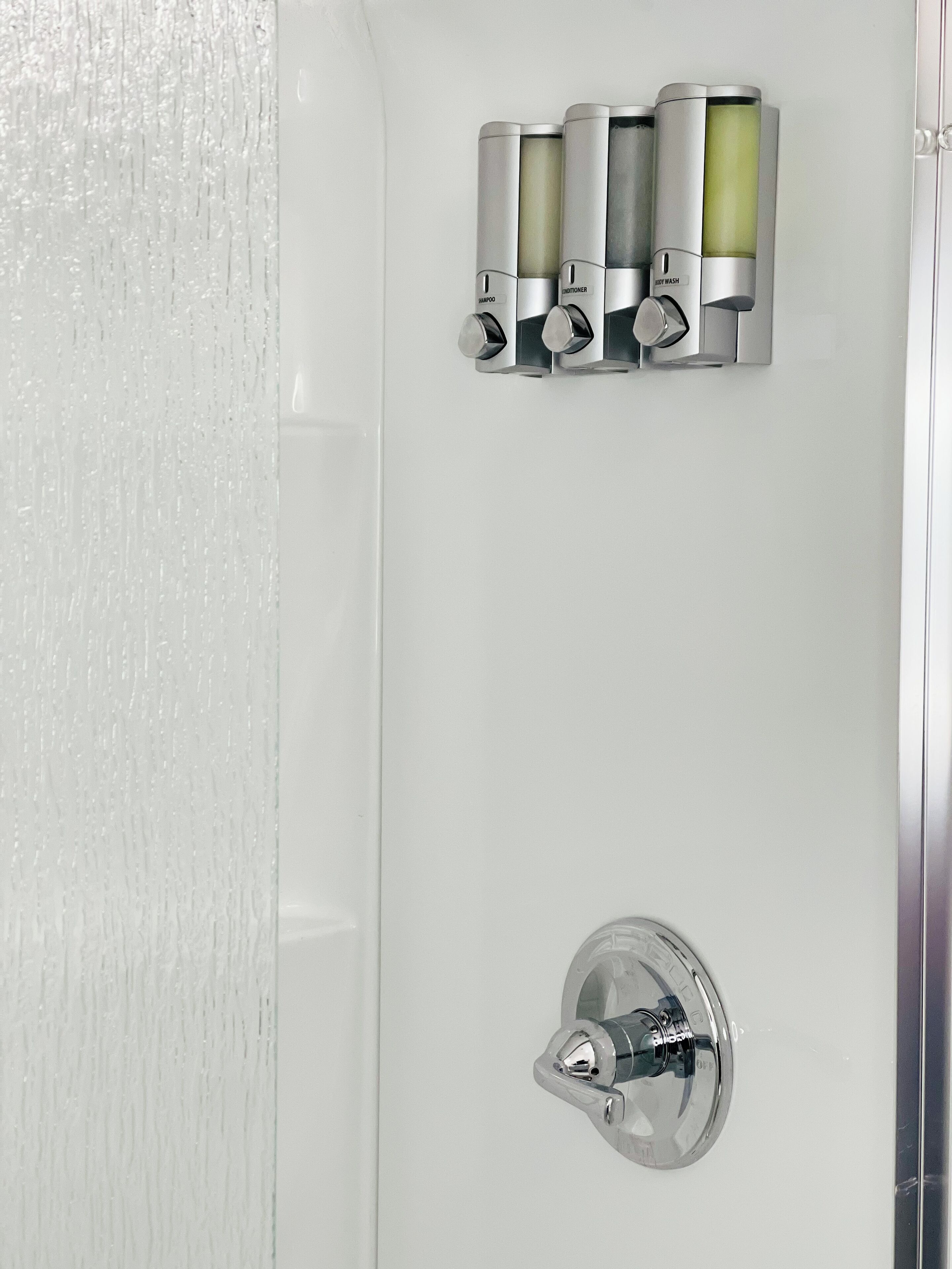 A bathroom with a white wall, a chrome faucet, and three bottles labeled shampoo, conditioner, and body wash.