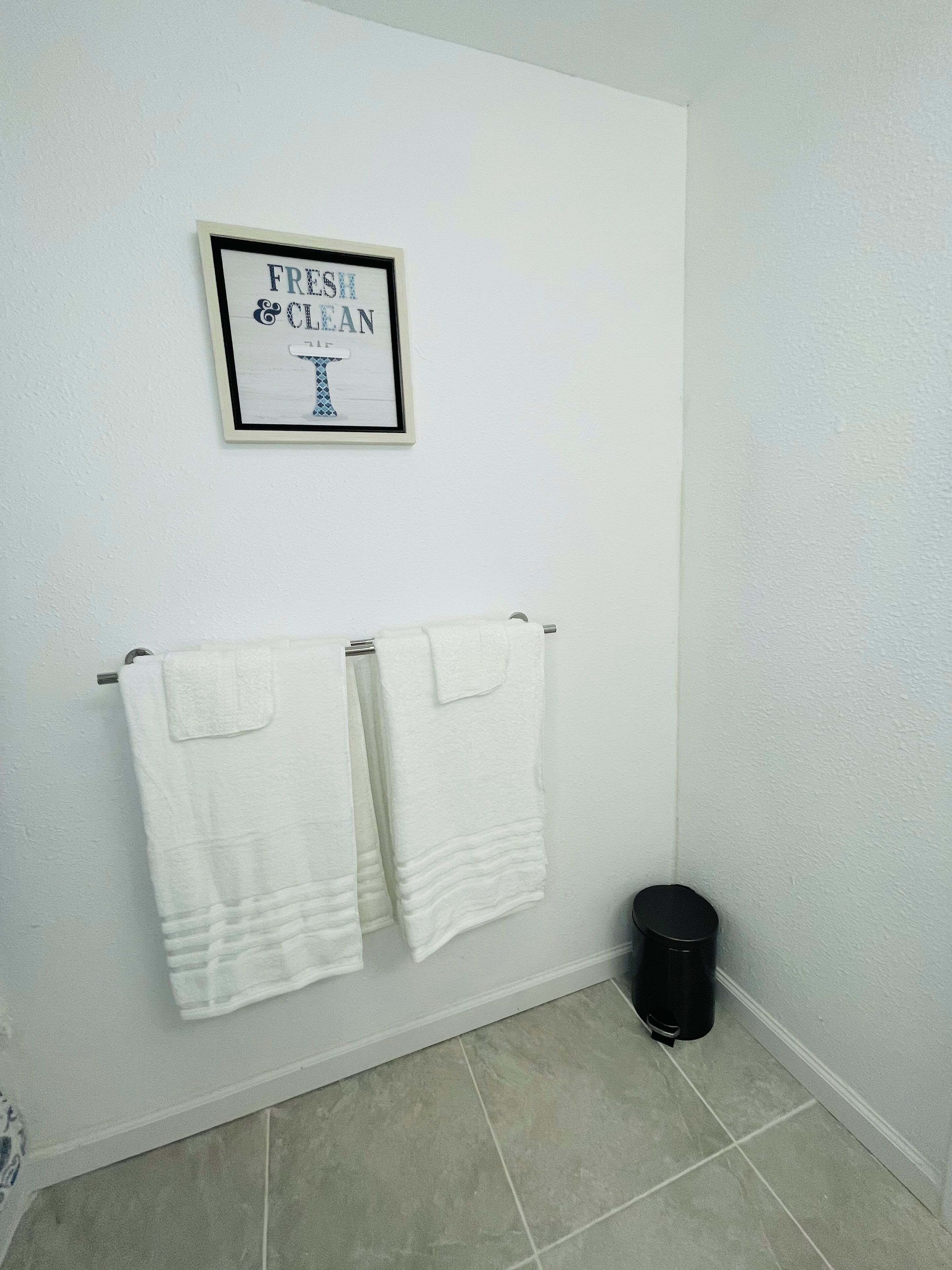 A bathroom with two white towels hanging on a rack, a black trash bin, and a framed sign above the towels that reads "FRESH CLEAN".