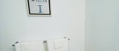A bathroom with two white towels hanging on a rack, a black trash bin, and a framed sign above the towels that reads "FRESH CLEAN".