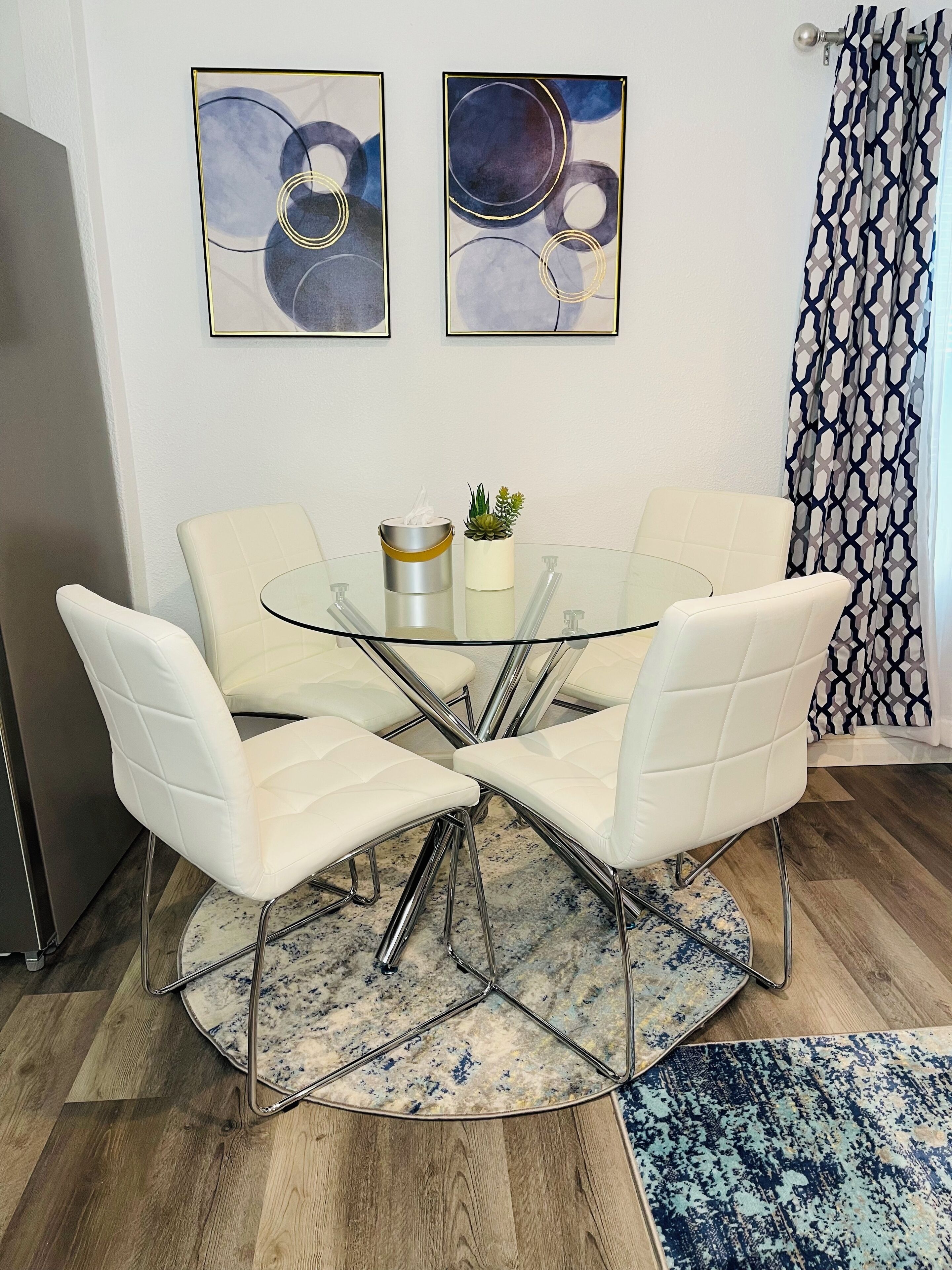 Een ronde glazen tafel met vier witte, beklede stoelen, een kleine potplant en een zakdoos. Op de muur hangen abstracte schilderijen en er is een gordijn met een patroon.