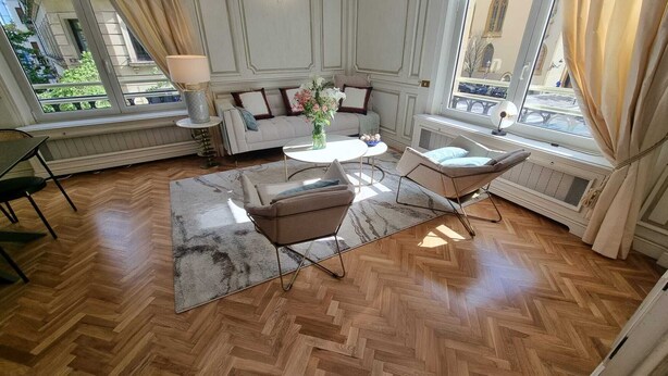 Une pièce spacieuse dotée d’un parquet en bois à motifs en chevrons, d’un canapé, de deux fauteuils, d’une petite table ronde et d’une fenêtre avec des rideaux.