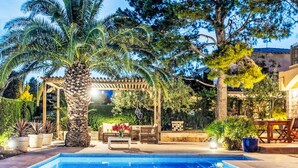 Ein Schwimmbecken mit einer Pergola und einer Sitzgelegenheit, umgeben von Pflanzen und Bäumen.
