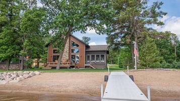 Une cabane moderne, dotée de fondations en pierre et de grandes fenêtres, située au bord d’un lac, avec une jetée en bois qui s’avance sur l’eau.
