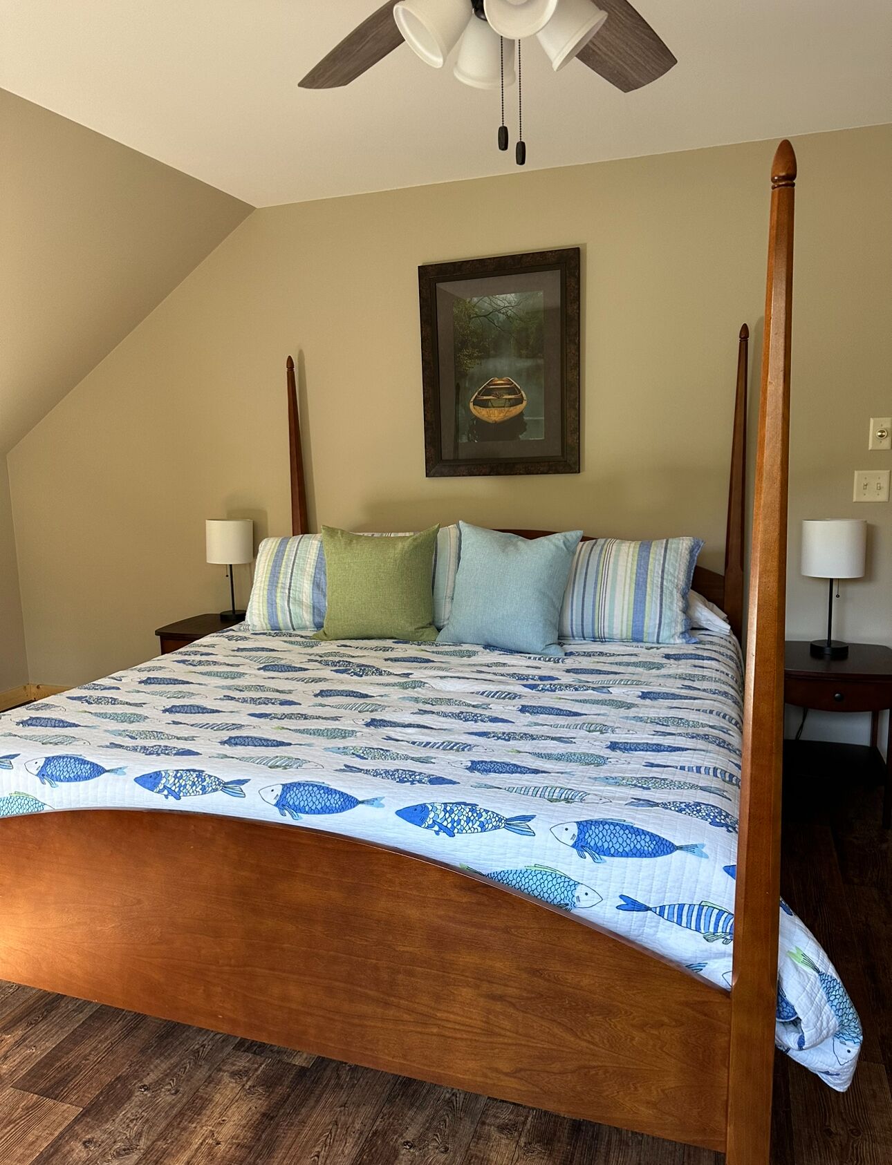 A four-poster bed with a fish-patterned bedspread, a framed picture on the wall, and a ceiling fan.