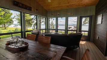 Une terrasse en bois avec une table et des chaises, de grandes fenêtres donnant sur un lac et un canapé en osier.