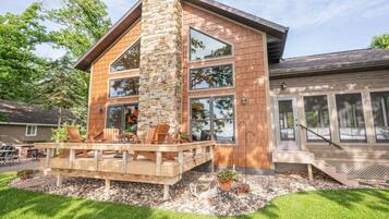 Une maison moderne à deux étages, dotée d’une cheminée en pierre, de grandes fenêtres et d’une terrasse en bois aménagée avec du mobilier d’extérieur.