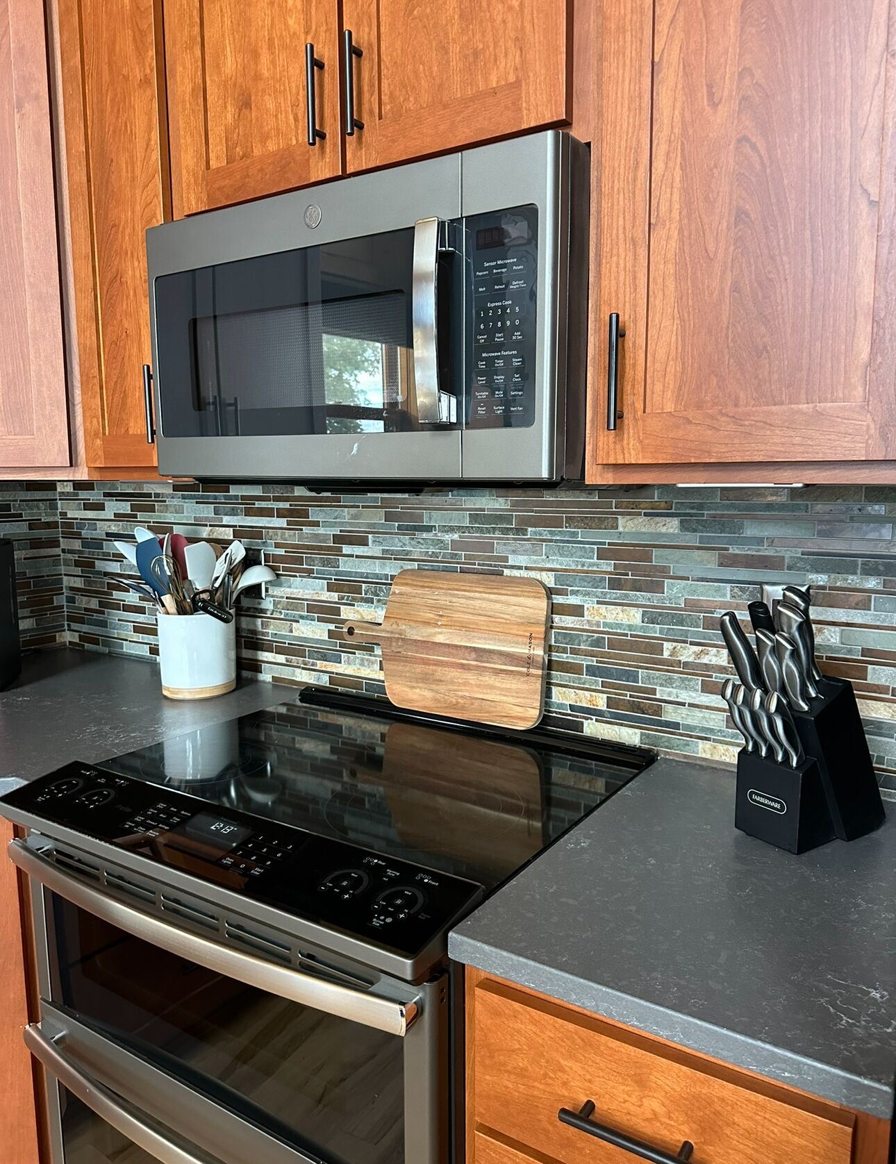 A kitchen with a stainless steel microwave, a wooden cutting board, and a knife block on the countertop.