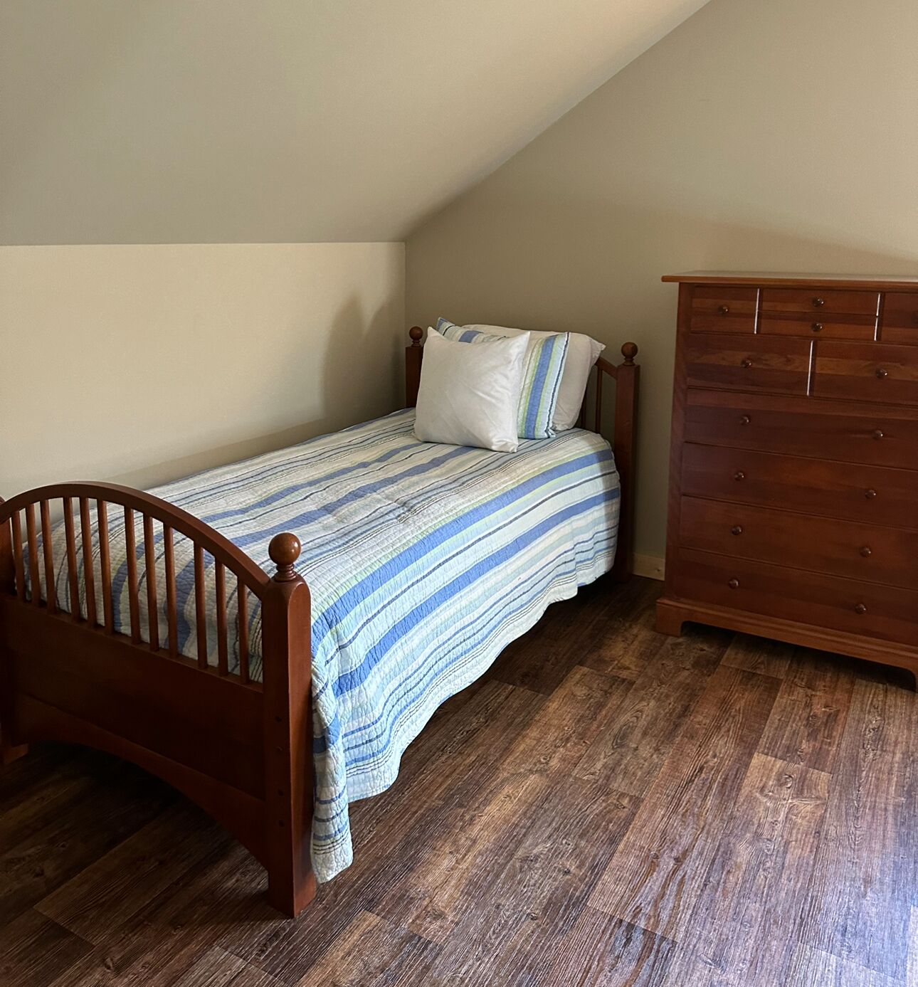 A small, single-bed room with a wooden headboard and a striped bedspread. There is a wooden dresser to the right and a sloped ceiling.