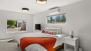 Une chambre moderne avec un grand lit, une télévision, une table de chevet avec une lampe, une fenêtre donnant sur des arbres et un appareil de climatisation.